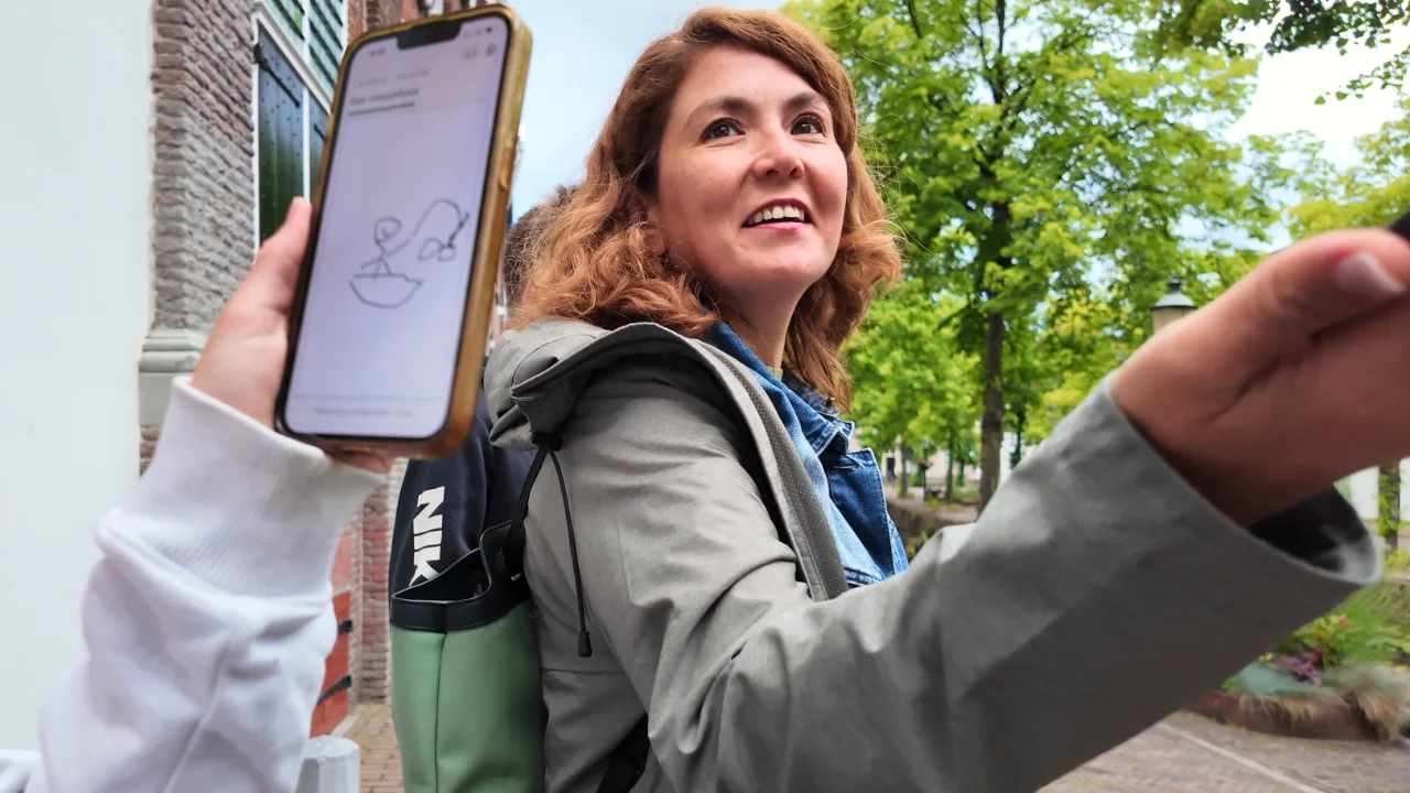 Lachend kijkt een vrouw omhoog in de oude binnenstad, terwijl iemand ernaast een telefoon ophoudt met een tekening uit het tekenspel